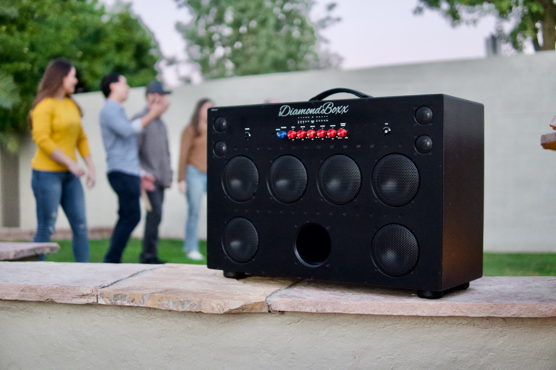 angled front view of black XL3 speaker with red and blue knobs in outdoor setting with people in background