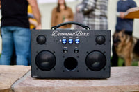 front view of black M3 speaker with blue knobs in outdoor setting with people around
