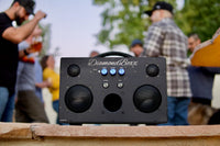 front view of black M3 speaker with blue knobs in outdoor setting with people around